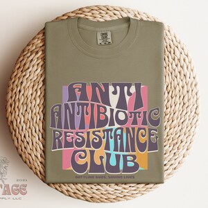 May include: A green t-shirt with a retro-style graphic that says "Antibiotic Resistance Club" in colorful letters. The text is surrounded by a pink, orange, and blue background. The bottom of the shirt says "Battling Bugs, Saving Lives".