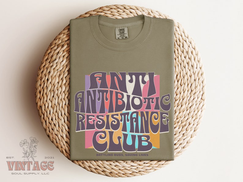 May include: A green t-shirt with a retro-style graphic that says "Antibiotic Resistance Club" in colorful letters. The text is surrounded by a pink, orange, and blue background. The bottom of the shirt says "Battling Bugs, Saving Lives".