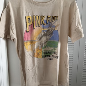Könnte beinhalten: Ein beiges Kurzarm-T-Shirt mit einem Grafikdruck von zwei mechanischen Händen, die sich schütteln. Der Text lautet PINK FLOYD, WISH YOU WERE HERE und 1975. Das kreisförmige Design enthält gelbe, rosa, blaue und grüne Farben auf dem beigen Stoff.