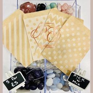 Puede incluir: Vista aérea de un recipiente transparente lleno de piedras preciosas pulidas y pequeñas bolsas de papel. Las bolsas tienen patrones de rayas, lunares y chevrones. El texto "Crystal Boutique" es visible en las bolsas.