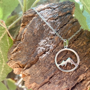 Puede incluir: Un collar de plata con un colgante circular que presenta una silueta de montaña.