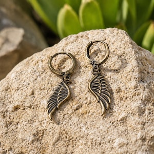 May include: A pair of bronze-toned earrings. Each earring has a small hoop and a wing-shaped charm with detailed feathering. The earrings are displayed on a textured, light-coloured rock, with green foliage in the background.