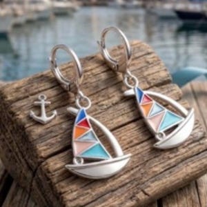 Puede incluir: Pendientes de plata con forma de velero y velas de esmalte de colores. Los pendientes presentan un diseño de velero con esmalte rojo, naranja, rosa, azul y blanco. También hay un charm de ancla. Los pendientes se exhiben sobre una superficie de madera envejecida.