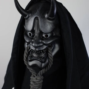 May include: A black Oni mask with curved horns and a menacing expression. The mask is detailed with sharp teeth and is connected to a thick, grey rope. The mask is set against a black hooded garment.