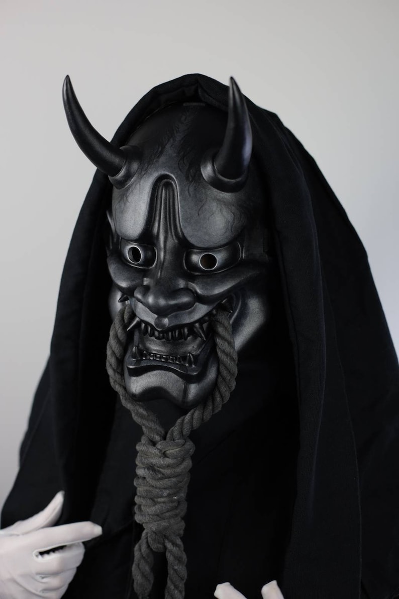May include: A black Oni mask with curved horns and a menacing expression. The mask is detailed with sharp teeth and is connected to a thick, grey rope. The mask is set against a black hooded garment.