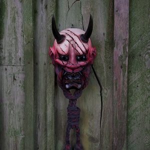 May include: A Japanese demon mask in red and white, featuring black horns and a fierce expression. The mask has scratch marks and is suspended by a dark rope with red detailing. It hangs against a weathered, green wooden backdrop.