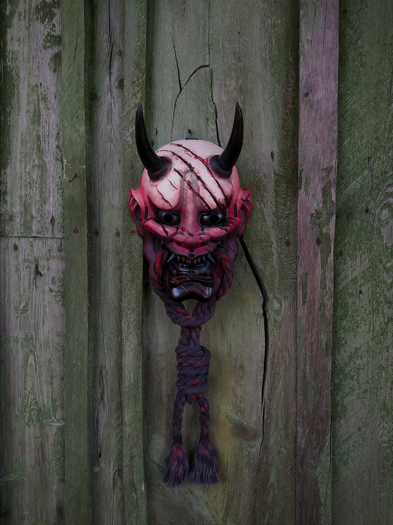 May include: A Japanese demon mask in red and white, featuring black horns and a fierce expression. The mask has scratch marks and is suspended by a dark rope with red detailing. It hangs against a weathered, green wooden backdrop.