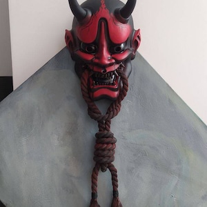 May include: A red and black Japanese Oni mask with black horns and a menacing expression. A thick, brown rope is tied around the mask's mouth and hangs down. The mask is mounted on a grey, diamond-shaped surface.