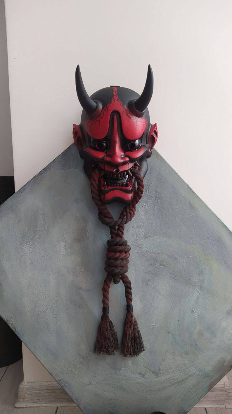 May include: A red and black Japanese Oni mask with black horns and a menacing expression. A thick, brown rope is tied around the mask's mouth and hangs down. The mask is mounted on a grey, diamond-shaped surface.