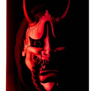 May include: A red and white Japanese Hannya mask with two large horns and a menacing expression. The mask features a detailed design with visible teeth and a rope-like element hanging from the mouth. The background is dark, emphasizing the mask's features.