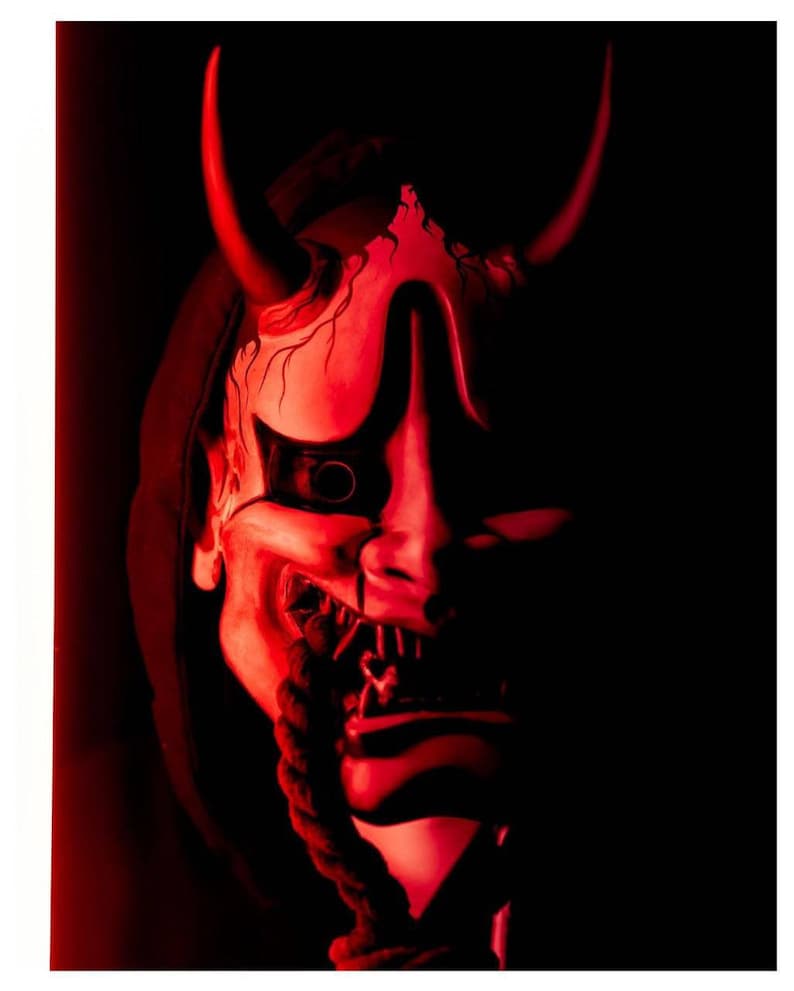 May include: A red and white Japanese Hannya mask with two large horns and a menacing expression. The mask features a detailed design with visible teeth and a rope-like element hanging from the mouth. The background is dark, emphasizing the mask's features.