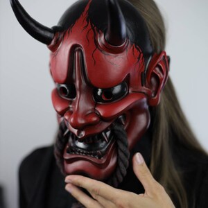 May include: A red and black Japanese demon mask with horns and a fierce expression. The mask has intricate details, including sharp teeth and a rope-like beard. The mask is a striking piece of art.