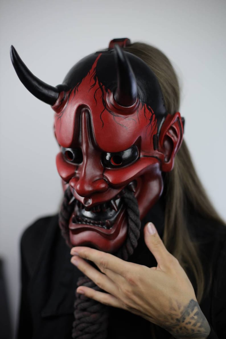 May include: A red and black Japanese demon mask with horns and a fierce expression. The mask has intricate details, including sharp teeth and a rope-like beard. The mask is a striking piece of art.