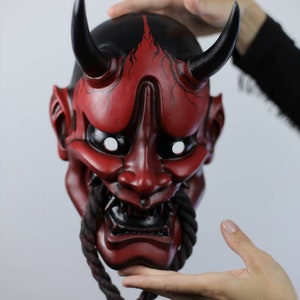 May include: A red and black Oni mask with curved horns and a rope beard. The mask has white eye sockets and a fierce expression. The top of the mask is black with red accents. The mask is held up by hands.