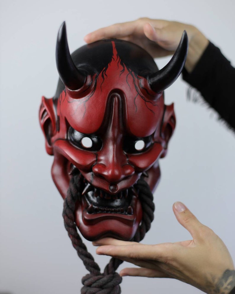 May include: A red and black Oni mask with curved horns and a rope beard. The mask has white eye sockets and a fierce expression. The top of the mask is black with red accents. The mask is held up by hands.