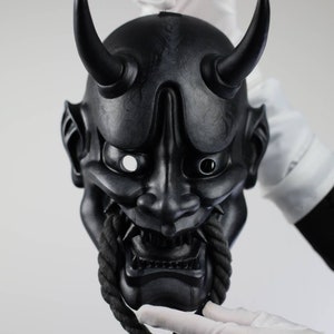 May include: A black Oni mask with two curved horns, a fierce expression, and a rope detail. The mask is held by gloved hands against a white background. The mask has sharp teeth and detailed facial features.