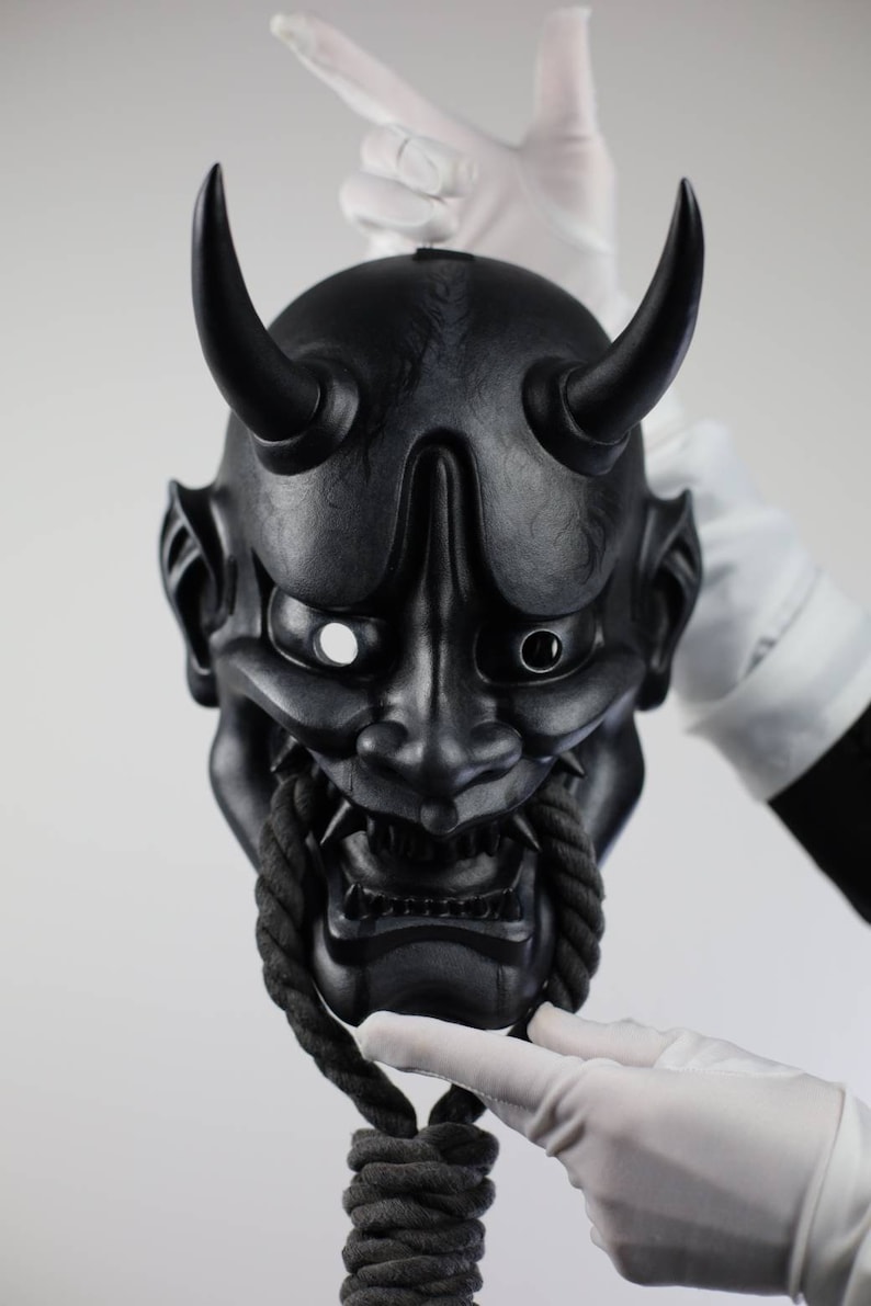 May include: A black Oni mask with two curved horns, a fierce expression, and a rope detail. The mask is held by gloved hands against a white background. The mask has sharp teeth and detailed facial features.