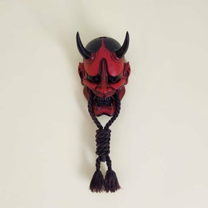 May include: A red and black Japanese Oni mask with curved horns and a fierce expression. The mask has a dark rope hanging from the bottom with two tassels. The mask is mounted on a light cream wall.