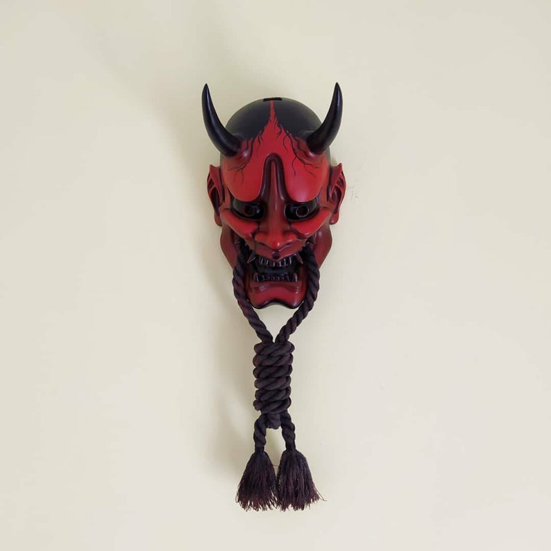 May include: A red and black Japanese Oni mask with curved horns and a fierce expression. The mask has a dark rope hanging from the bottom with two tassels. The mask is mounted on a light cream wall.