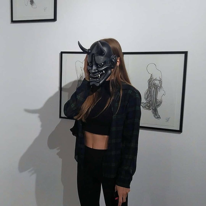 May include: A black Oni mask with horns and a menacing expression. The mask is held up in front of a person's face. The person is wearing a black crop top, black leggings, and a green and black plaid shirt. Artwork is visible on the wall.