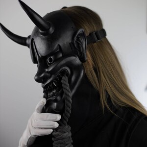 May include: A black Oni mask with two large horns, sharp teeth, and a menacing expression. A thick, dark rope hangs from the mask's mouth. The mask is worn with a black strap and a black outfit. A white gloved hand touches the rope.