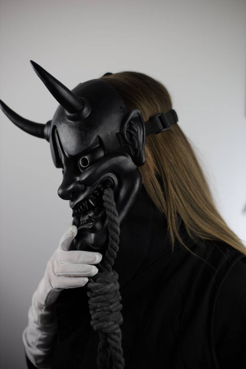 May include: A black Oni mask with two large horns, sharp teeth, and a menacing expression. A thick, dark rope hangs from the mask's mouth. The mask is worn with a black strap and a black outfit. A white gloved hand touches the rope.