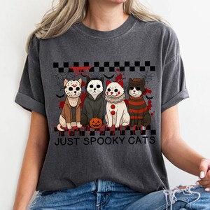 May include: Dark gray t-shirt featuring four cartoon cats dressed as horror movie characters. The cats are arranged in a row with a Halloween theme, and the text "JUST SPOOKY CATS" is printed below them.