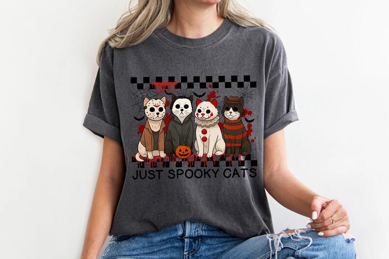 May include: Dark gray t-shirt featuring four cartoon cats dressed as horror movie characters. The cats are arranged in a row with a Halloween theme, and the text "JUST SPOOKY CATS" is printed below them.