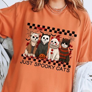 May include: Orange t-shirt with a graphic of four cats dressed as horror movie characters. The cats are in costumes, including Jason Voorhees, Michael Myers, a clown, and Freddy Krueger. The text "JUST SPOOKY CATS" is below the cats.