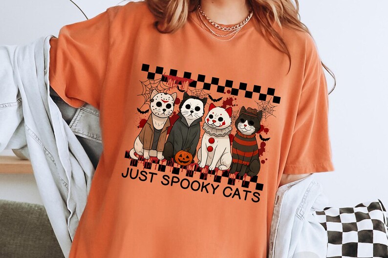 May include: Orange t-shirt with a graphic of four cats dressed as horror movie characters. The cats are in costumes, including Jason Voorhees, Michael Myers, a clown, and Freddy Krueger. The text "JUST SPOOKY CATS" is below the cats.