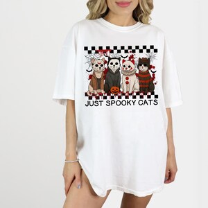May include: White t-shirt featuring a graphic of four cats dressed in Halloween costumes. The cats are arranged above the text "JUST SPOOKY CATS" on a black and white checkered pattern. The design includes a pumpkin.