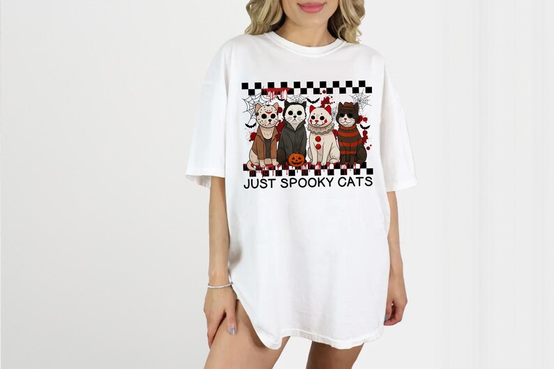 May include: White t-shirt featuring a graphic of four cats dressed in Halloween costumes. The cats are arranged above the text "JUST SPOOKY CATS" on a black and white checkered pattern. The design includes a pumpkin.