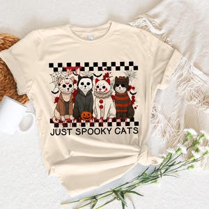 May include: Cream-colored t-shirt featuring a graphic of four cats dressed in Halloween costumes. The cats are arranged in a row, with a pumpkin in front of them. The text "JUST SPOOKY CATS" is below the cats.