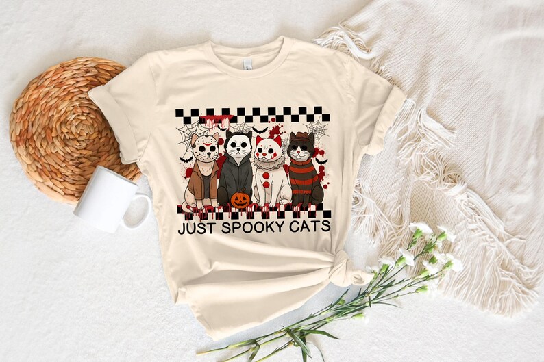 May include: Cream-colored t-shirt featuring a graphic of four cats dressed in Halloween costumes. The cats are arranged in a row, with a pumpkin in front of them. The text "JUST SPOOKY CATS" is below the cats.