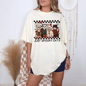 May include: Cream-colored t-shirt featuring a graphic of four cats dressed as horror movie characters, with a pumpkin and spiderwebs. The text "JUST SPOOKY CATS" is below the graphic. The shirt is worn by a person.