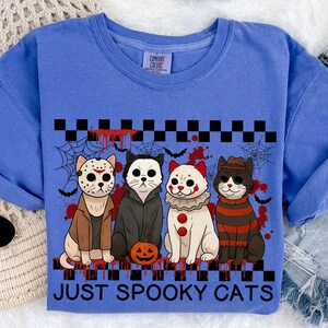 May include: A blue Comfort Colors t-shirt featuring four cartoon cats dressed in Halloween costumes. The cats are in costumes like Jason Voorhees, a vampire, a clown, and Freddy Krueger. The words "JUST SPOOKY CATS" are printed below the cats.