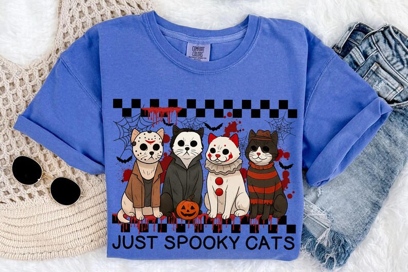 May include: A blue Comfort Colors t-shirt featuring four cartoon cats dressed in Halloween costumes. The cats are in costumes like Jason Voorhees, a vampire, a clown, and Freddy Krueger. The words "JUST SPOOKY CATS" are printed below the cats.