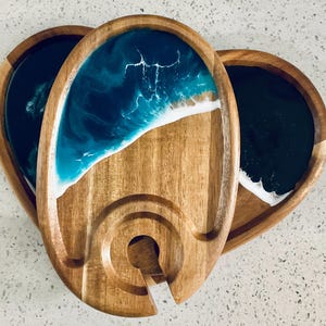 Puede incluir: Una bandeja de servir de madera con una incrustación de resina azul y blanca que representa una ola del océano. La bandeja tiene un hueco para una botella y es perfecta para servir queso, galletas y otros aperitivos.