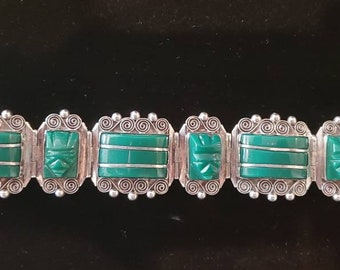 Op de afbeelding: Een zilveren armband met groene jade stenen en ingewikkelde ontwerpen. Elke steen is gezet in een vierkant frame met een gegraveerd gezicht in het midden.