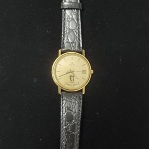 May include: A gold wristwatch with a black leather strap. The watch face is gold with black hands and markers. The strap has a crocodile skin pattern. The watch face has the text "Liberty 1886-1986".
