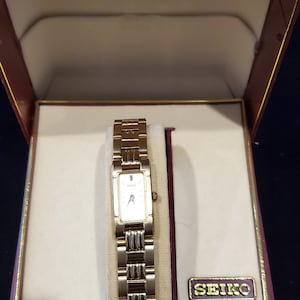 May include: A gold-tone Seiko watch with a rectangular face and a gold metal band. The watch is displayed in a burgundy box with a gold lining and the word "SEIKO" printed in gold on the box.