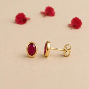 Puede incluir: Un par de pendientes de botón de oro con rubíes rojos facetados de forma ovalada en un engaste liso. Un pendiente se muestra de frente, mientras que el otro está colocado para mostrar el poste de oro y el mecanismo de cierre de mariposa.