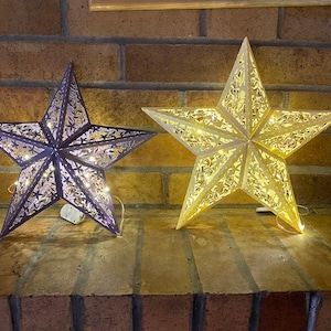 以下が含まれることがあります： レンガの壁に、薄い茶色と濃い紫色の装飾的な星型のライトが2つ吊り下げられています。ライトは暖色系の白色LEDで照らされています。