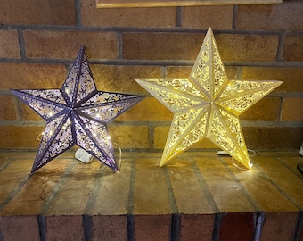 May include: Two decorative star-shaped lights, one in a light brown color and one in a dark purple color, are hanging on a brick wall. The lights are illuminated with warm white LED lights.