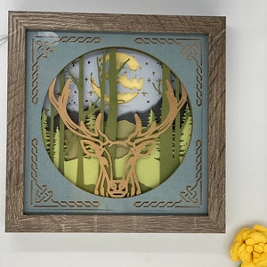 May include: A framed paper art piece with a deer silhouette in a forest scene. The deer is cut out of brown paper and the forest is cut out of green paper. The background is a blue sky with a yellow moon and stars.