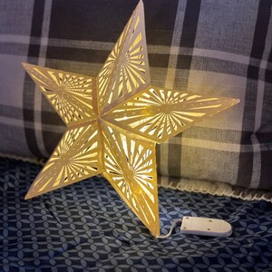 May include: A gold star-shaped paper lantern with a white cord and battery pack. The lantern is lit and casting a warm glow. The lantern is sitting on a blue and white patterned fabric.