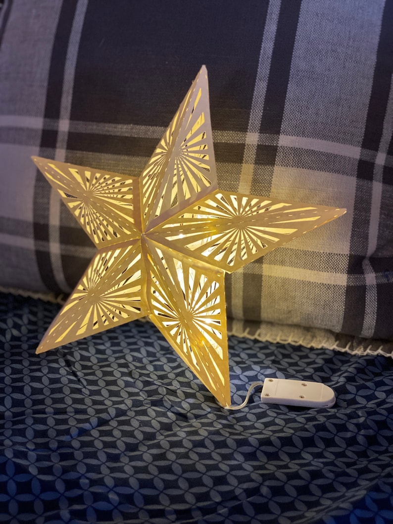 May include: A gold star-shaped paper lantern with a white cord and battery pack. The lantern is lit and casting a warm glow. The lantern is sitting on a blue and white patterned fabric.