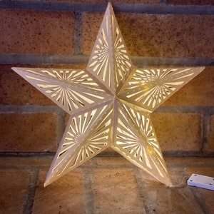 May include: A white, paper star with a cut-out design, illuminated from within by a string of lights. The star is hanging from a brick wall.