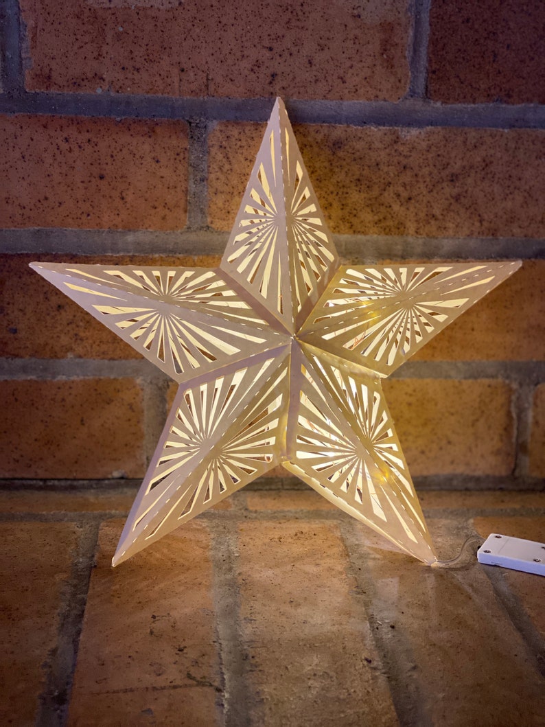 May include: A white, paper star with a cut-out design, illuminated from within by a string of lights. The star is hanging from a brick wall.