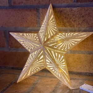 以下が含まれることがあります： 幾何学模様の白い紙の星で、中に電飾が施されています。星はレンガの壁に掛けられています。